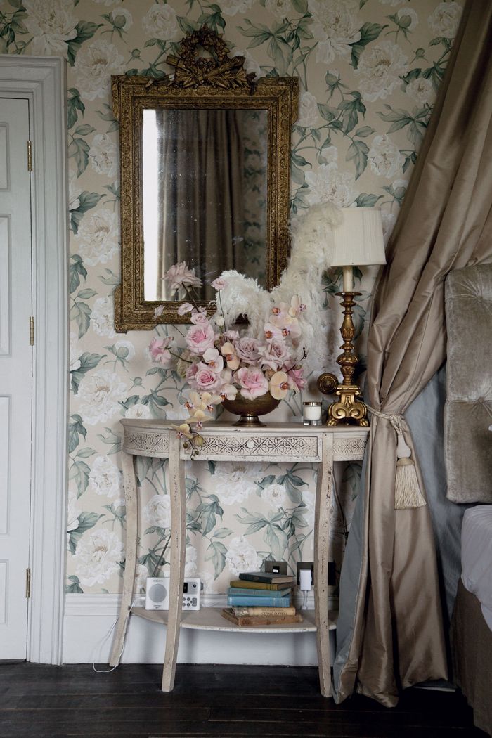 Entry corner with pale botanical wallpaper, a carved gilt mirror and a demilune console with flowers