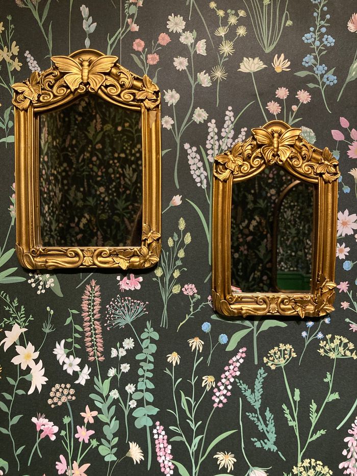 Two ornate gilt mirrors hung on dark charcoal wallpaper printed with wildflowers
