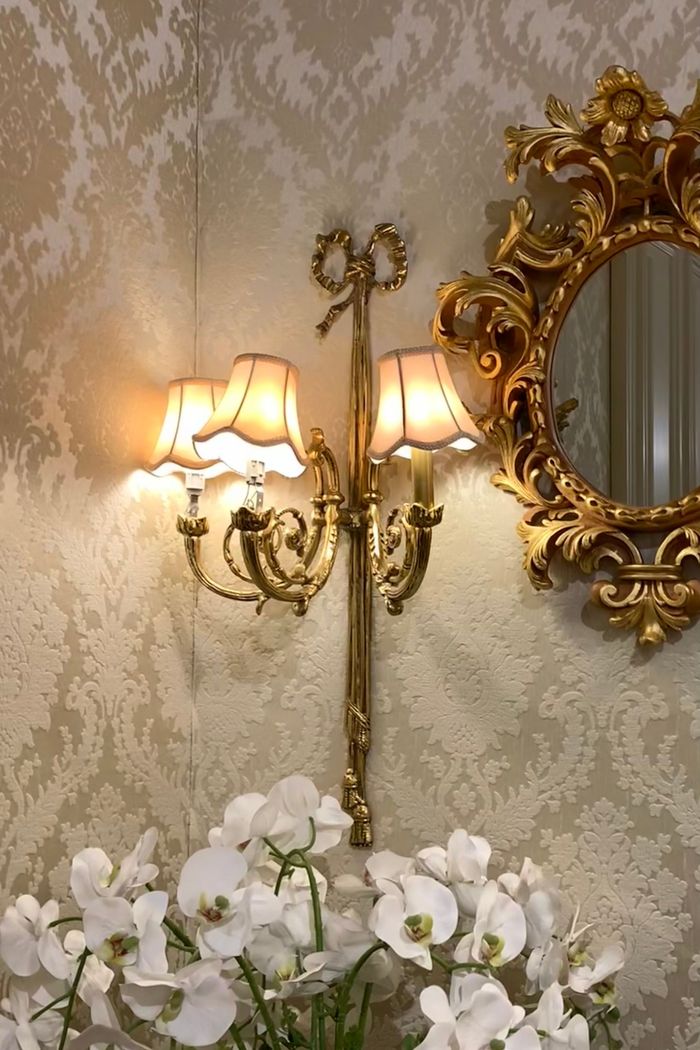 Gold sconce and rococo mirror on cream tone-on-tone damask wallpaper above white orchids