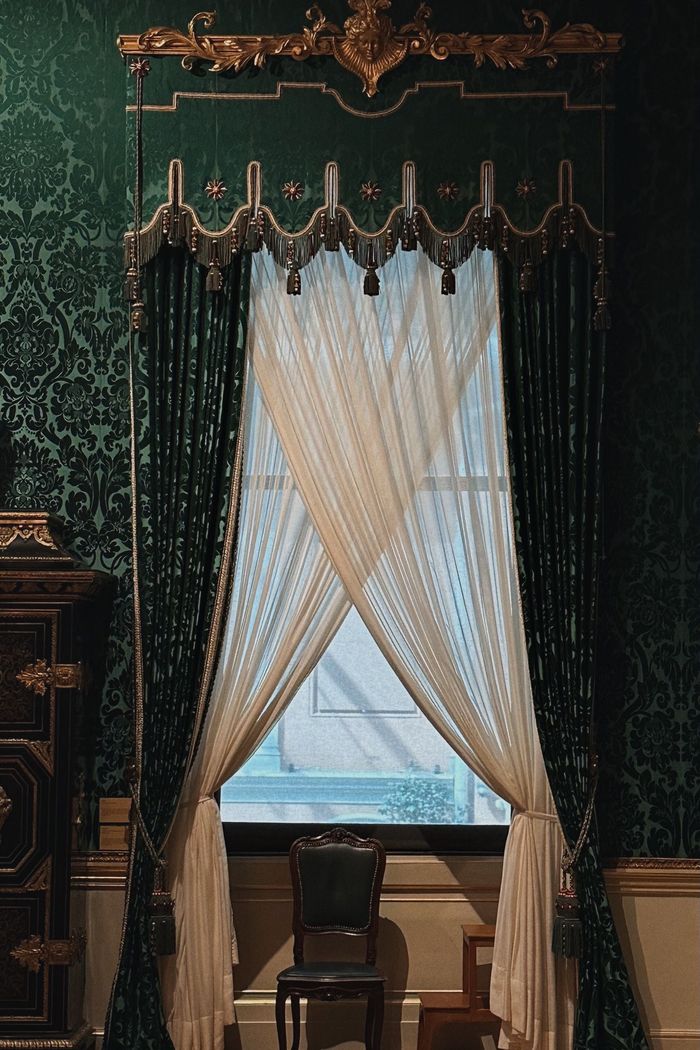Victorian-style room with deep green damask wallpaper framing a tall window dressed in sheer and green curtains