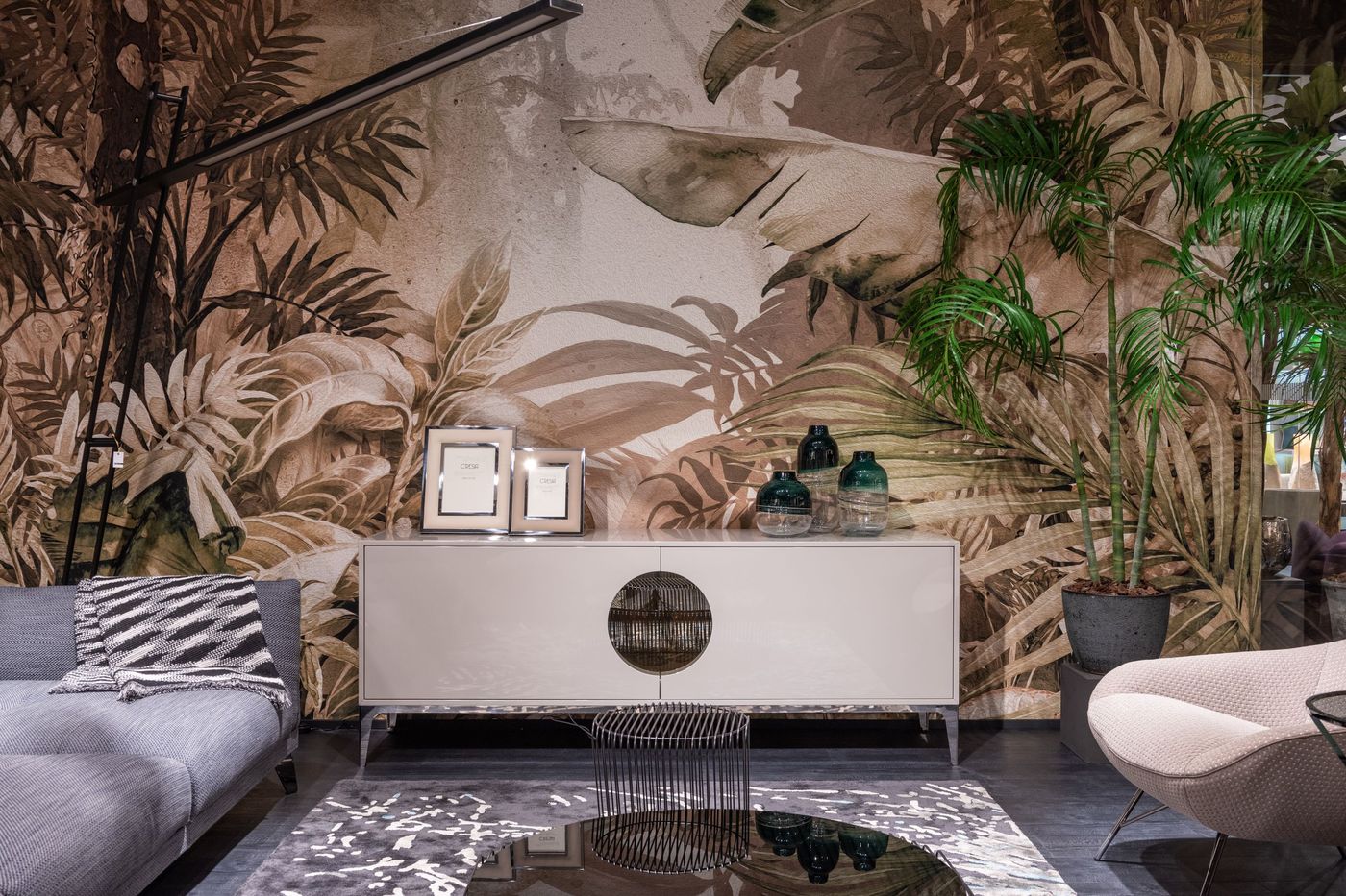 Living room with a full-wall tropical leaf mural wallpaper in muted sand and olive tones behind a pale console and grey sofa