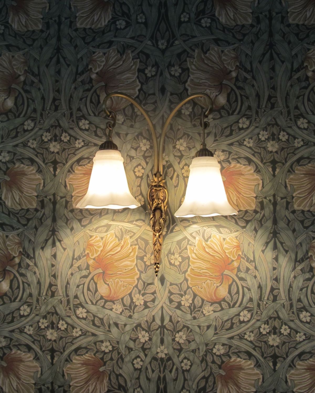 Brass twin wall sconce glowing against dark Arts and Crafts floral wallpaper