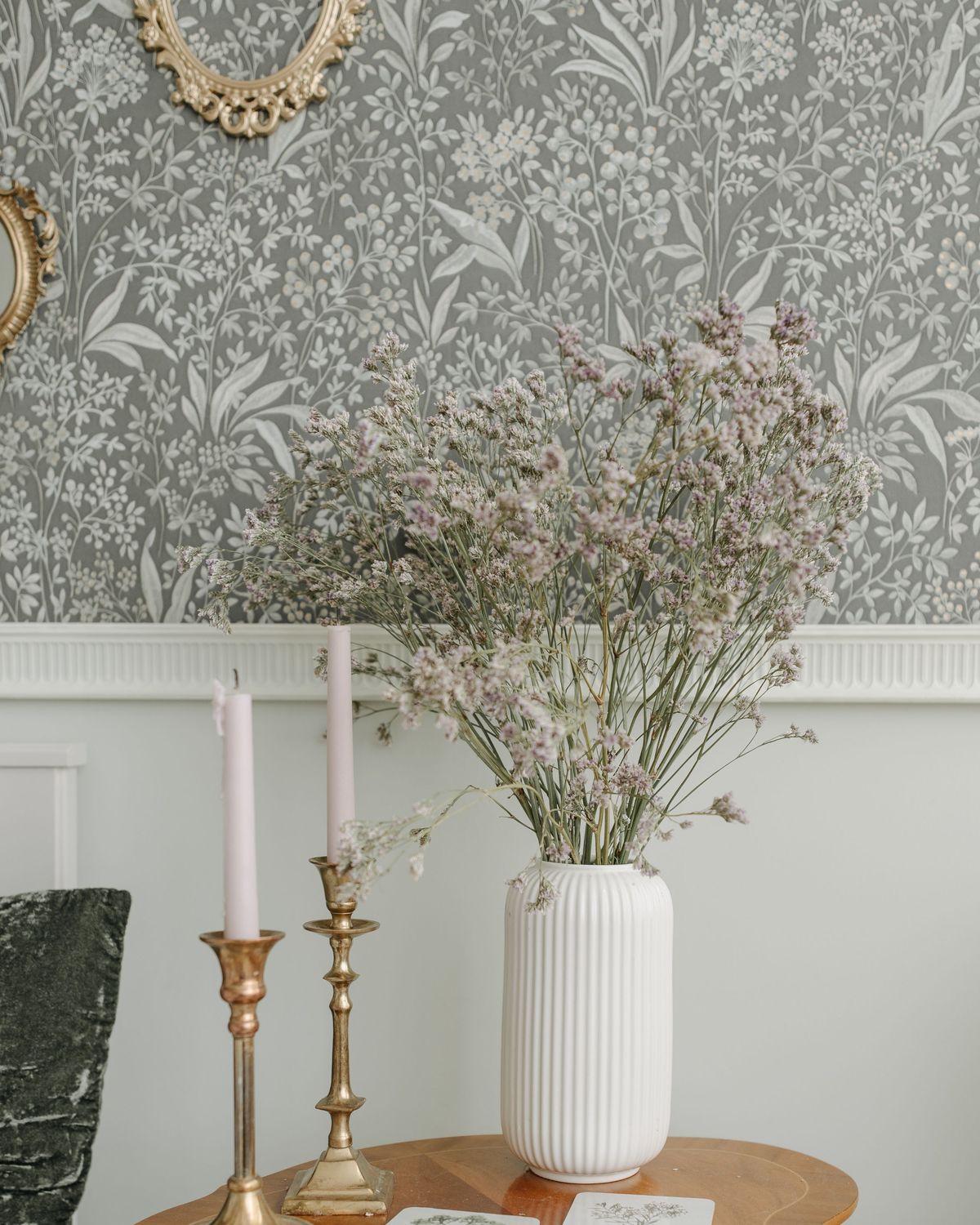 Grey botanical wallpaper above white wainscoting, with brass candlesticks and a vase of dried flowers on a round side table